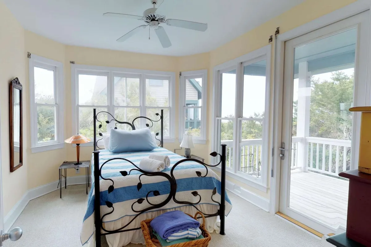 A cozy bedroom with a decorative iron bed frame sits against large windows that let in sunlight, accompanied by a basket of fresh towels and a softly glowing table lamp. The view beyond the window hints at the calming green of the surrounding maritime forest, reinforcing the tranquil and private retreat feeling described in the section.