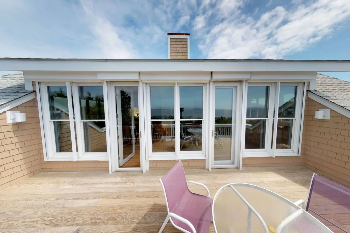 A spacious deck with comfortable chairs and large glass doors opens up to panoramic views of the ocean. The tranquil setting showcases a perfect spot for relaxing, group dinners, or entertaining friends, with the sunset casting a golden light over the space.