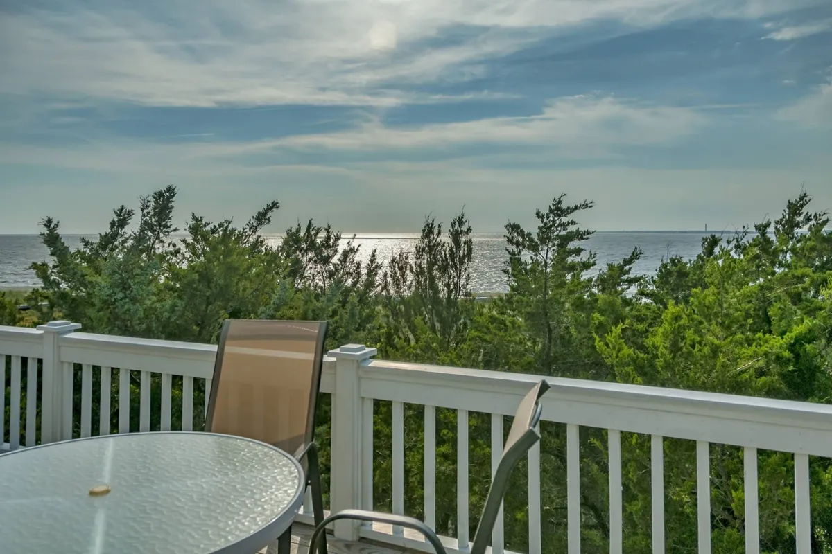 An outdoor dining table on a deck overlooks a sweeping ocean view, framed by lush greenery. The setting is peaceful and inviting, hinting at cozy dinners or relaxing evenings spent together above the shoreline.