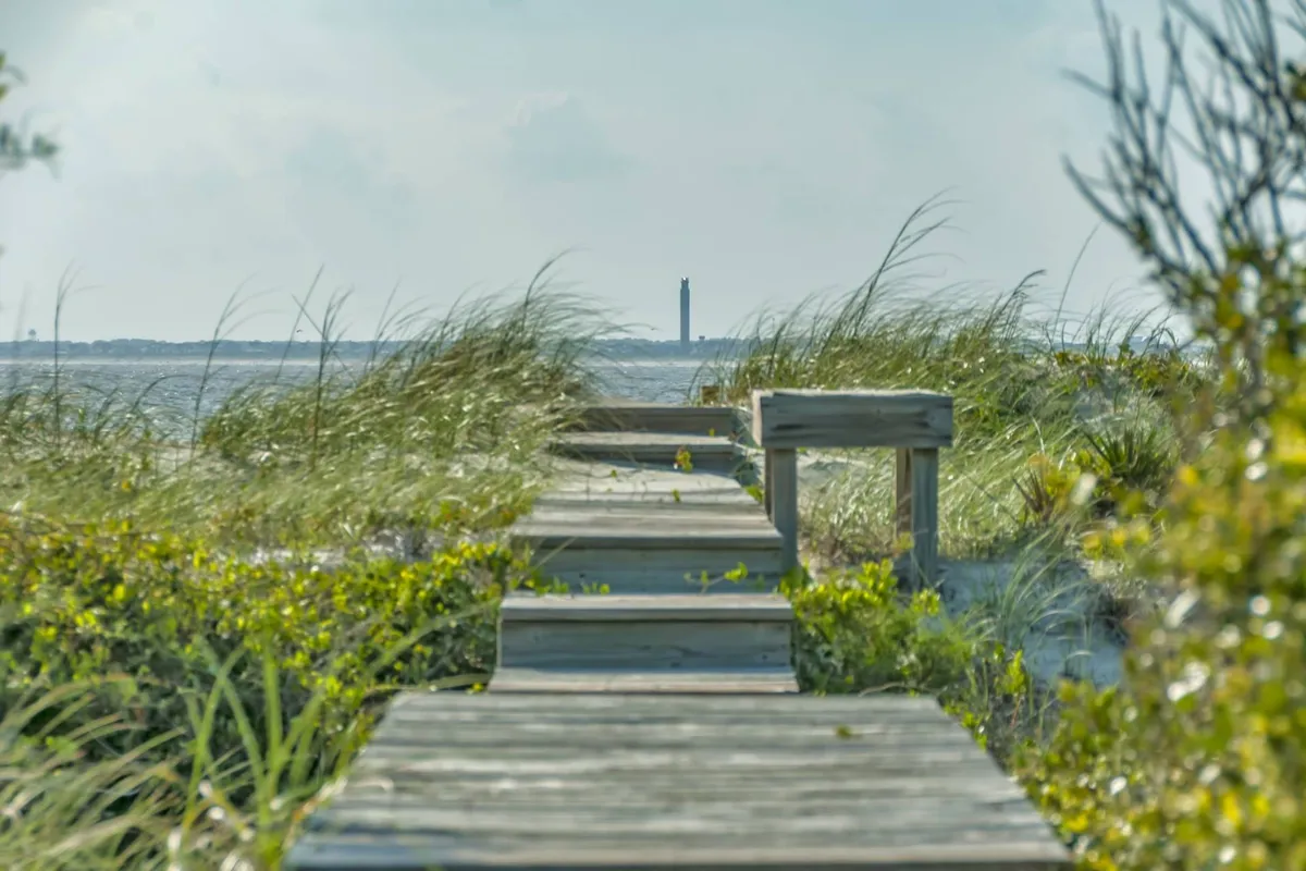 A serene wooden walkway stretches through dune grass toward the beach, with a distant lighthouse visible on the horizon. The scene is open, natural, and inviting, showcasing easy access to outdoor adventure and peaceful moments by the water.