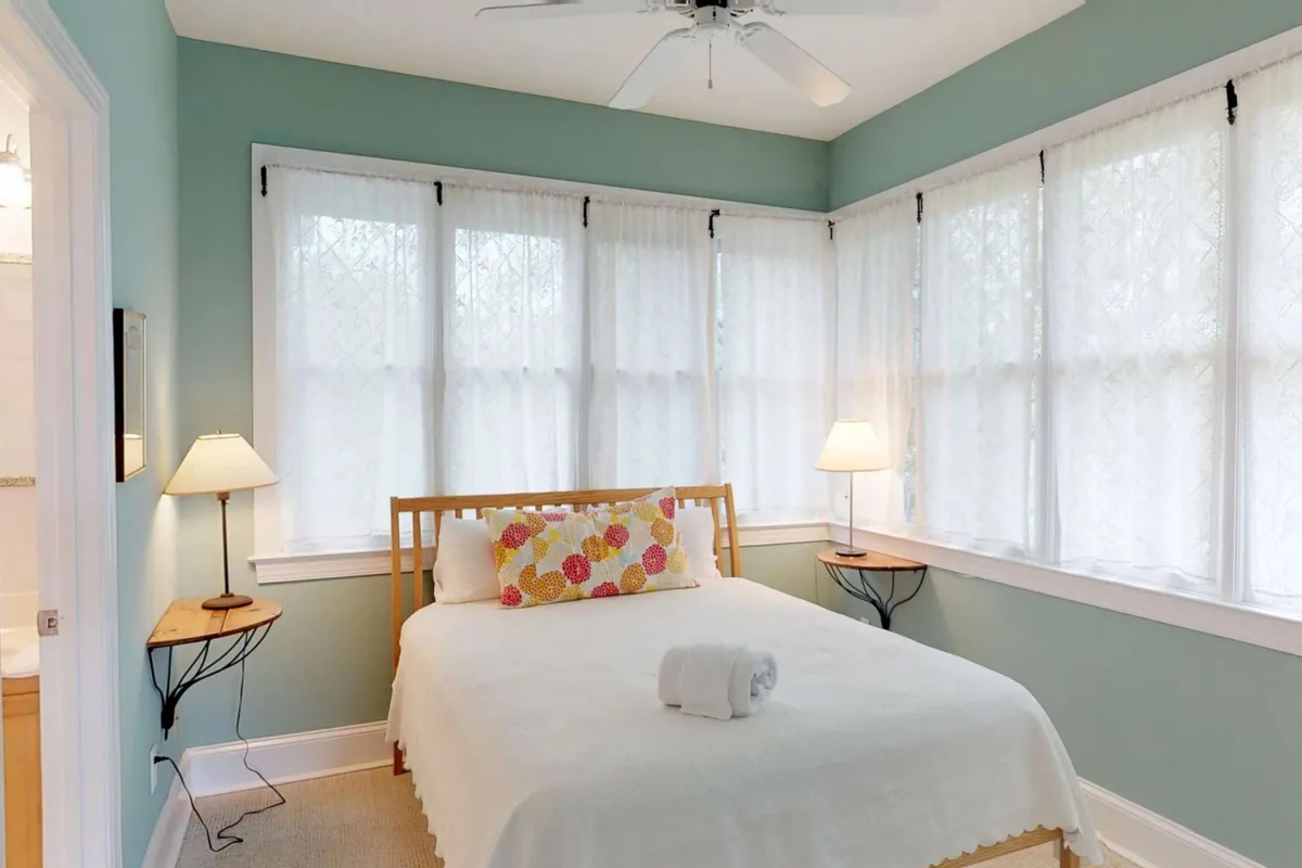 A bright, inviting bedroom featuring a cozy queen bed adorned with a decorative pillow. Large windows with sheer curtains bathe the room in soft, natural light, while bedside lamps create a welcoming ambiance ideal for relaxation after a day of adventure.