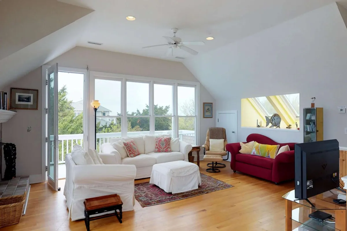 This image showcases a cozy living area with a comfortable white sofa, a red accent sofa, a coffee table, and large windows flooding the room with natural light. The setup is inviting, designed for relaxation and unwinding in a private home setting.