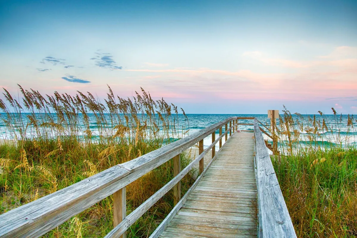 A wooden boardwalk winds through dune grass towards the ocean, bathed in the warm glow of sunset. The sky is soft and expansive, enhancing the tranquil, intimate atmosphere perfect for couples.