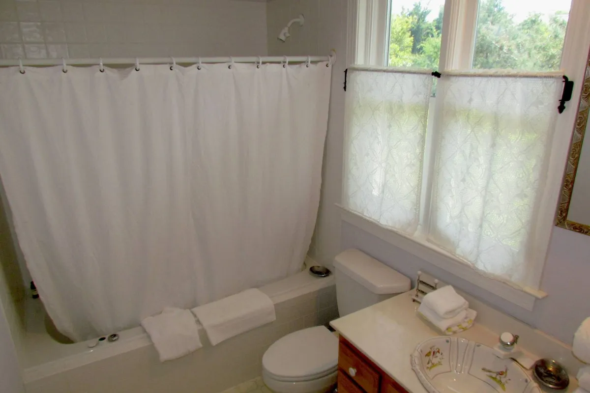 A tranquil bathroom with a bathtub and shower curtain, a vanity with a sink, and a window dressed in sheer curtains. The space feels airy and clean, ideal for unwinding and self-care, with sunlight streaming in to enhance relaxation.