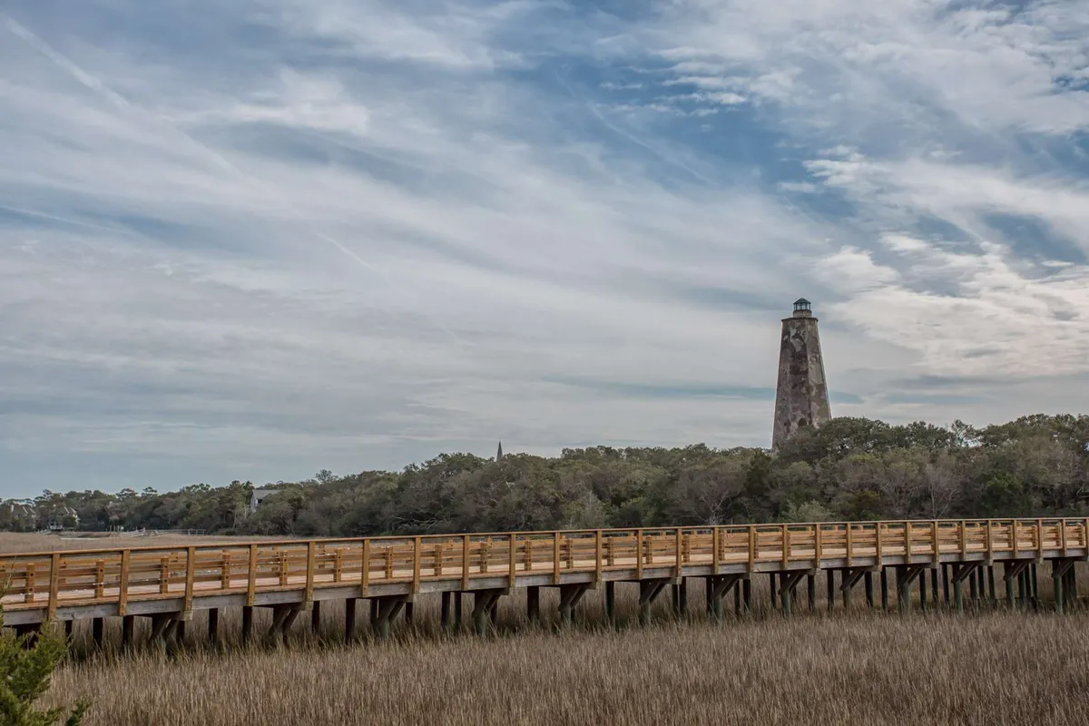 This image showcases the iconic Old Baldy Lighthouse standing tall near the marshland, with a wooden boardwalk stretching toward it. The tranquil surroundings and sunset ambiance evoke a sense of peaceful exploration and romantic strolls in nature.