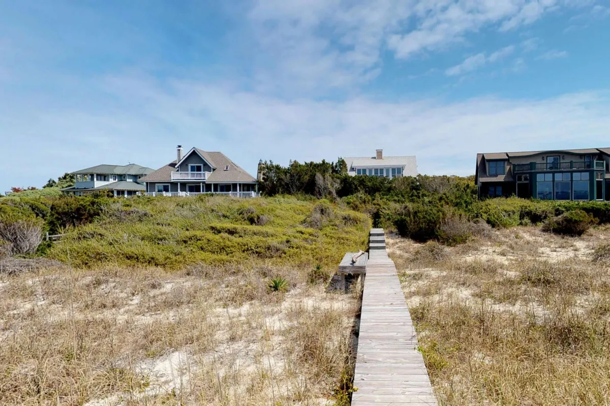 A boardwalk leads past natural vegetation and beach houses, creating a relaxed coastal atmosphere. The surroundings suggest a neighborhood with easy access to nature and scenic walks along the shore.
